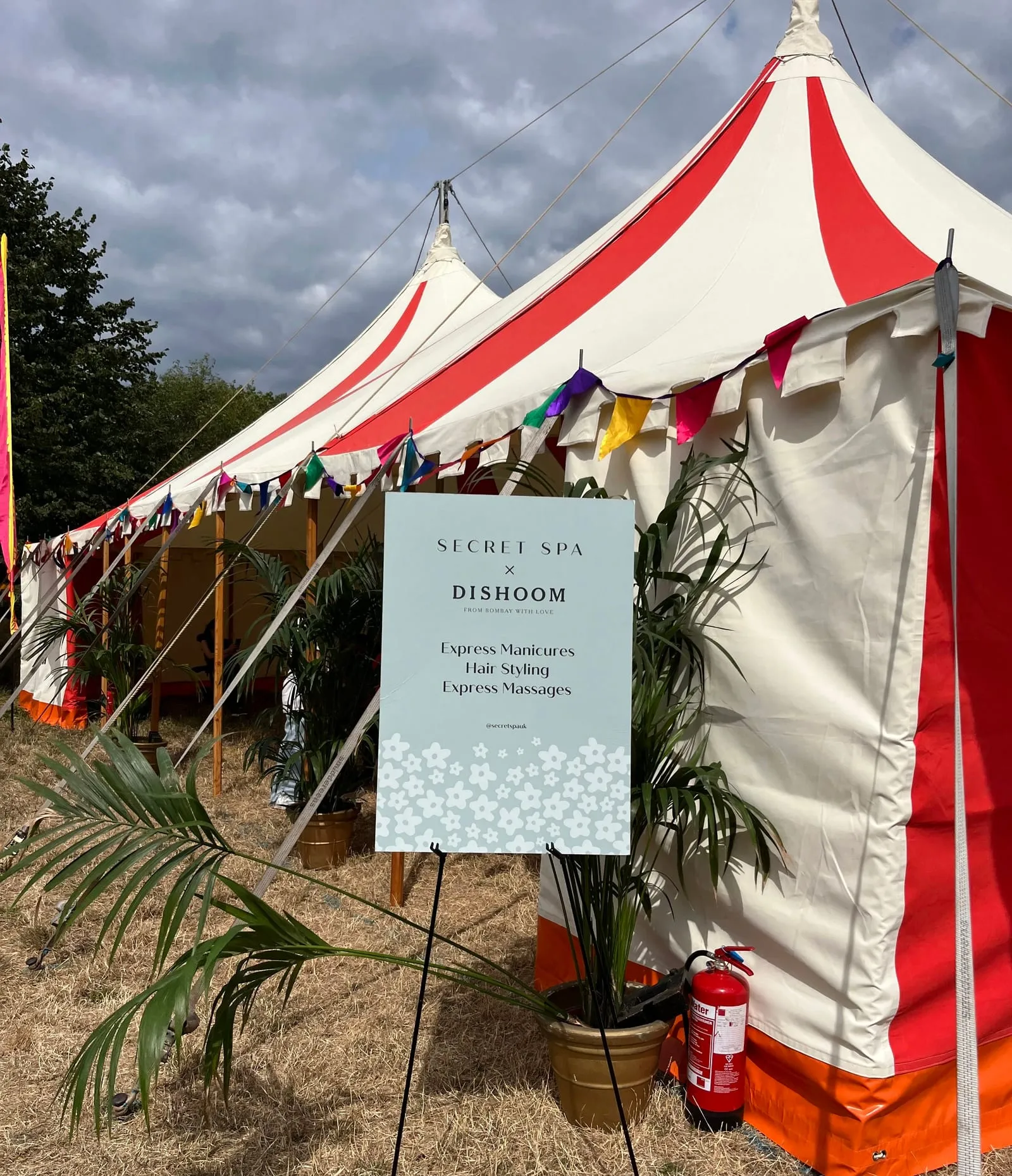 Secret Spa x Dishoom sign outside a red and white festival marquee, listing express manicures, hair styling and express massages