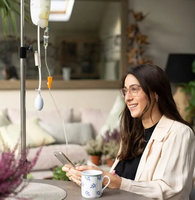 A client receiving an intravenous drip at home, seated beside the drip stand with a mug and phone