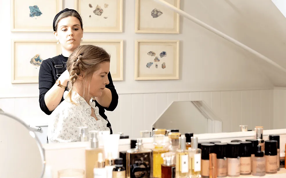 Hairdresser plaiting a clients hair at a dressing table crowded with skincare and perfume bottles