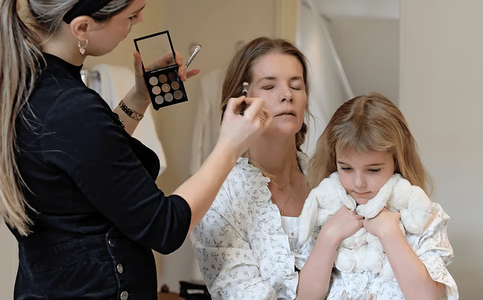 Make-up artist applying eyeshadow from a palette while a client holds her young daughter on her lap
