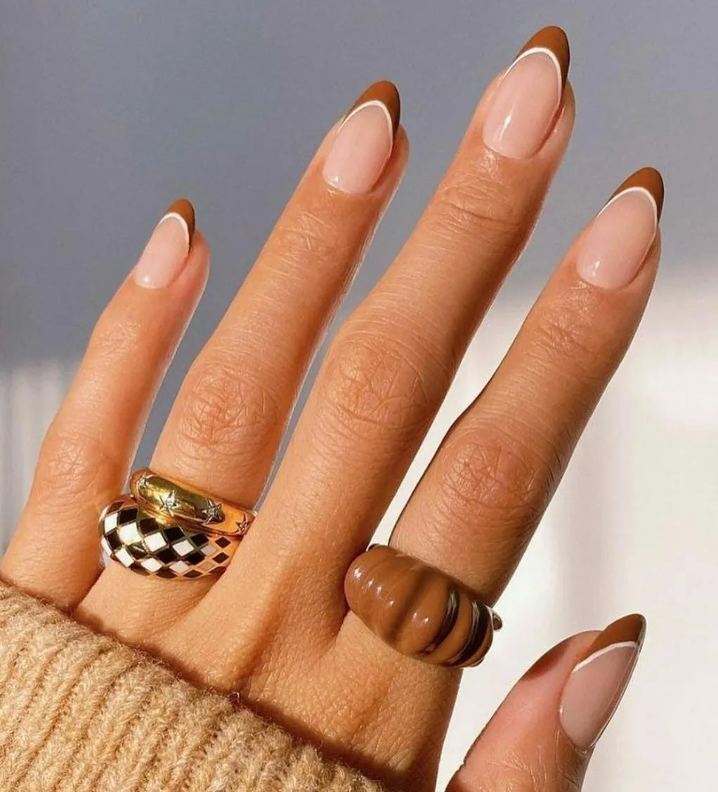 Almond nails with burnt orange French tips outlined in white, worn with chunky rings