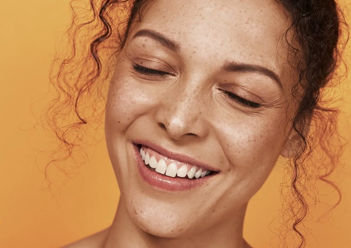 Close-up of a smiling face with freckles and clear, glowing skin against an orange backdrop