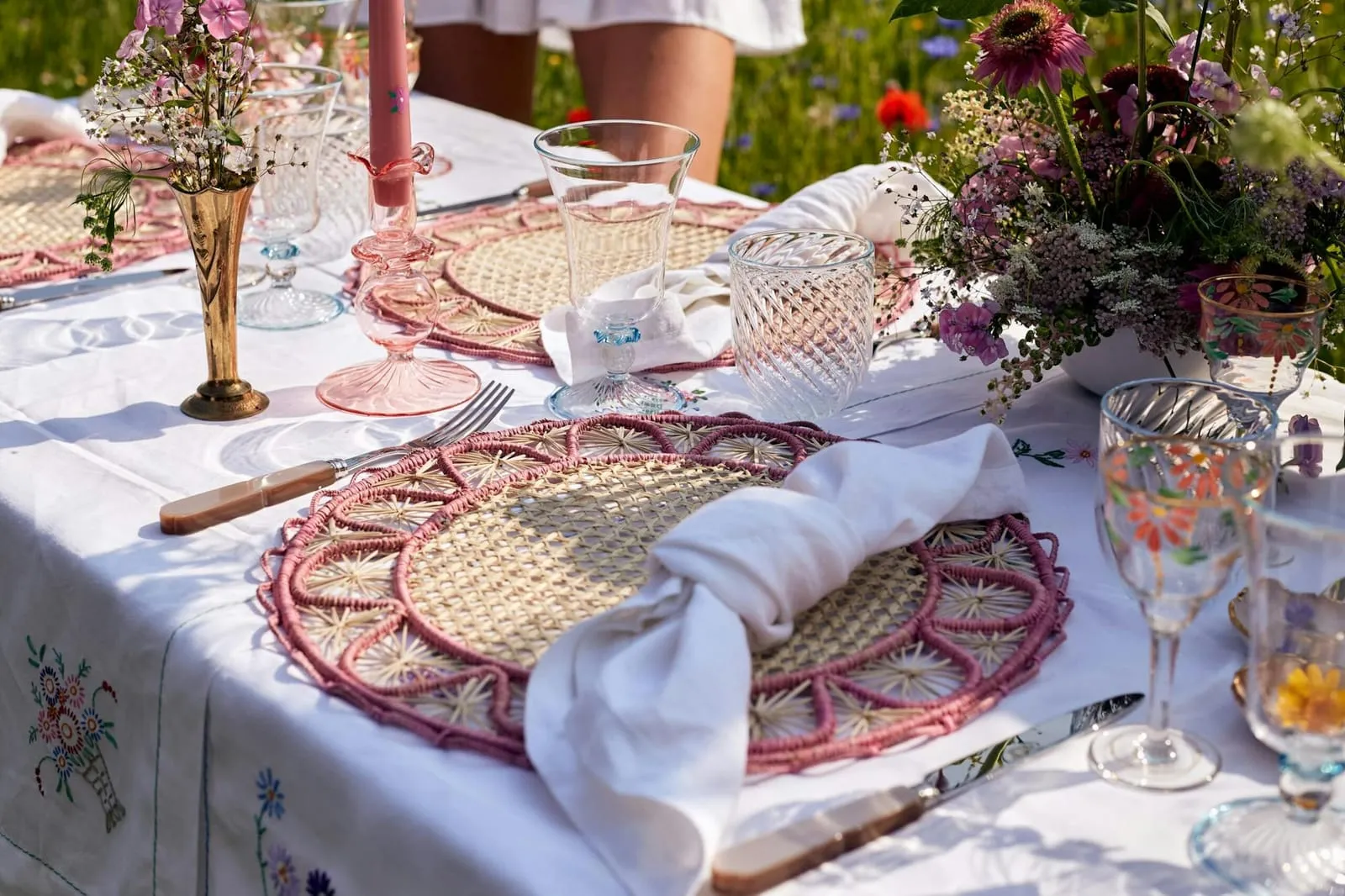 Sunlit outdoor tablescape with pink raffia placemats, embroidered linen, vintage glassware and wildflowers