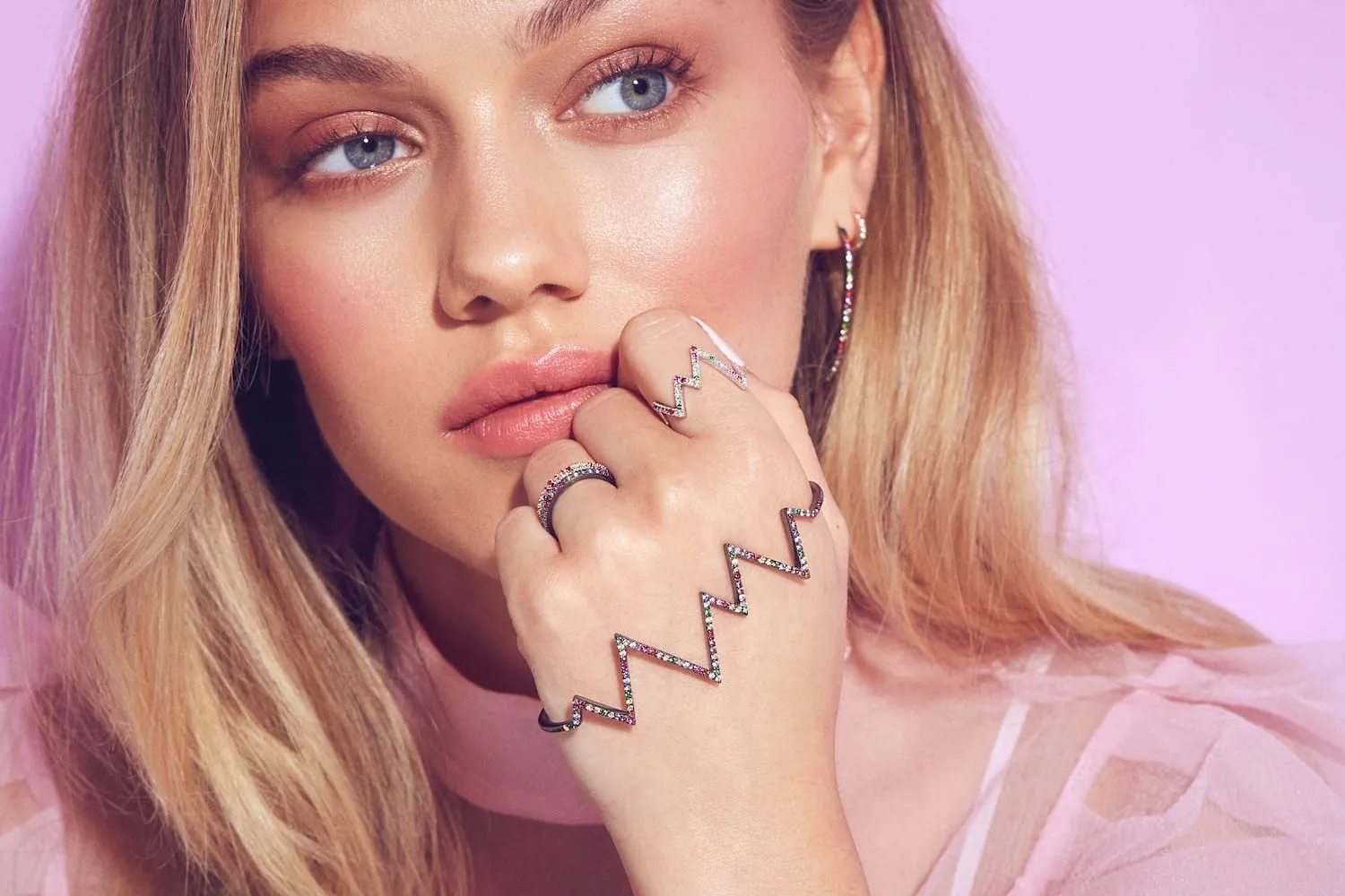 Close-up of glossy skin and nude lips with rainbow-gem zigzag rings held against the mouth