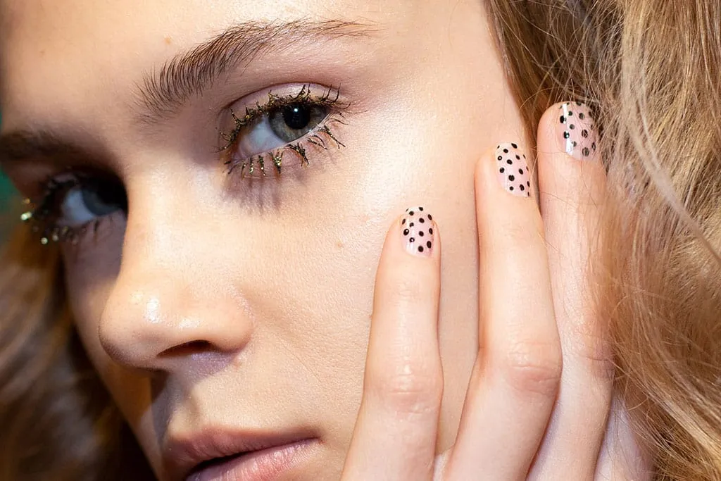Close-up of gold glitter-flecked lashes worn with sheer nude nails dotted in black