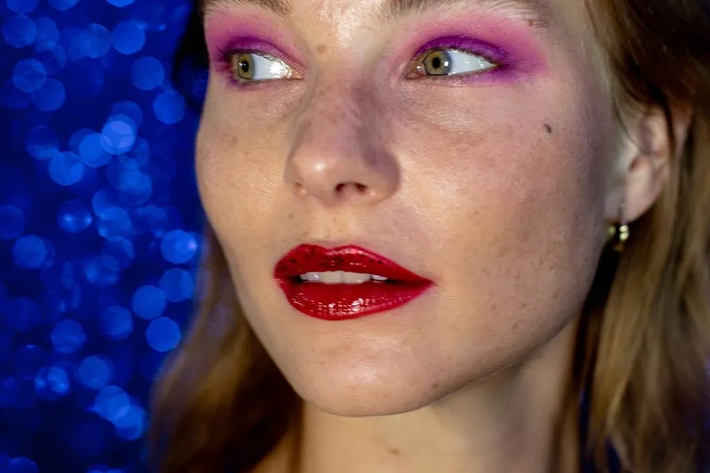 Glossy deep red overdrawn lip worn with pink and purple shadow washed across the lids