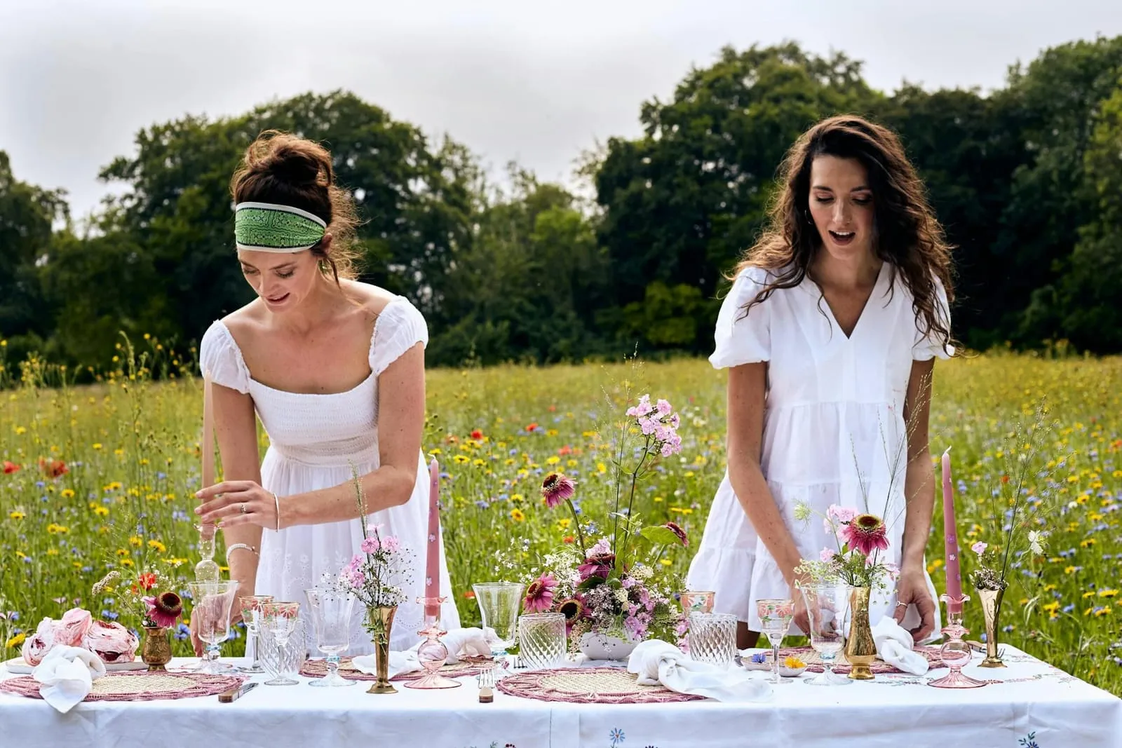 Two event stylists laying a candlelit table with pink wildflowers in a summer meadow