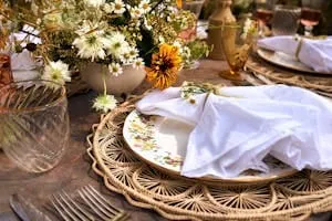 Rustic place setting layering a white napkin over a floral plate and woven charger, with wildflowers behind