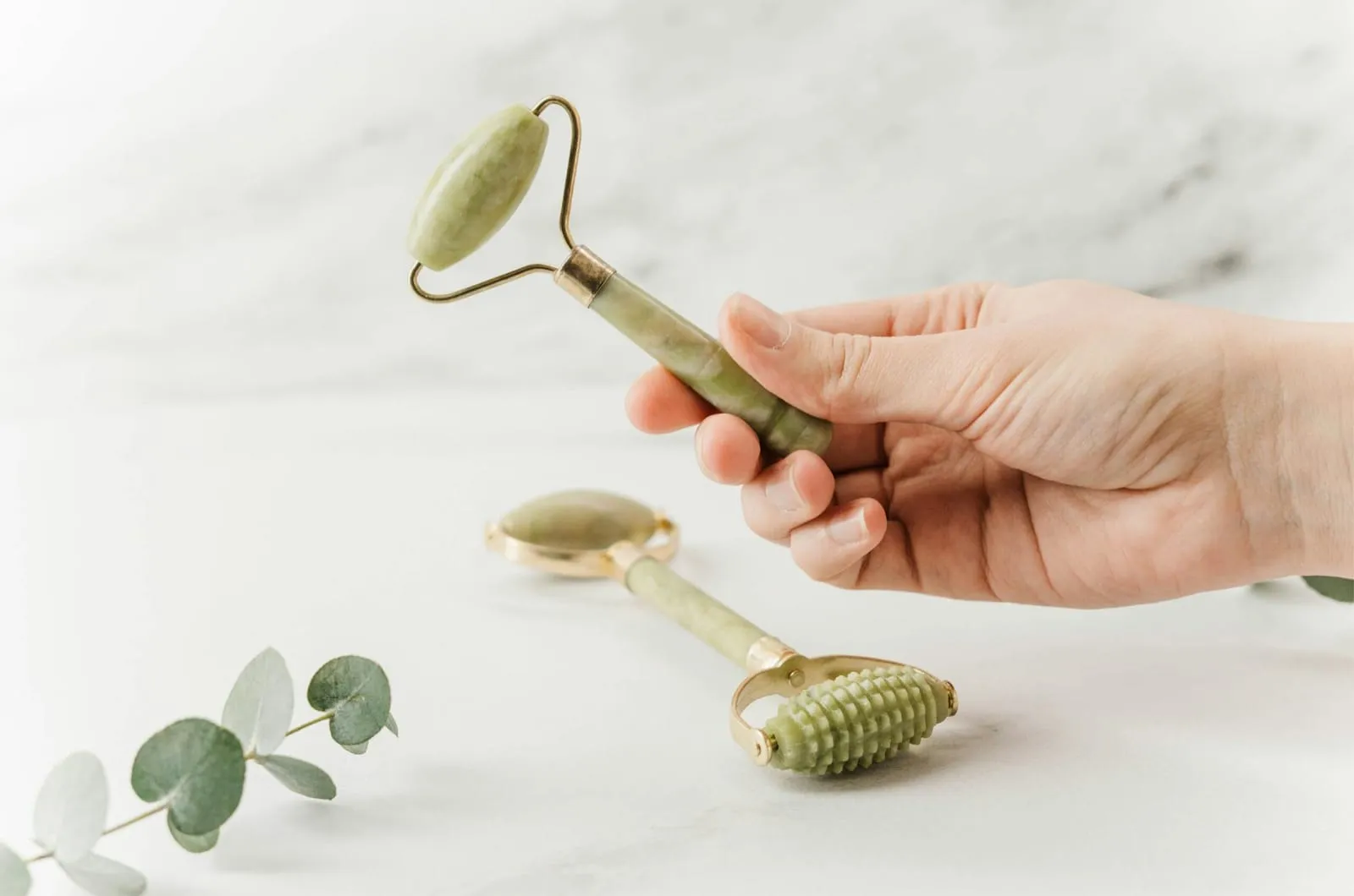 Hand holding a jade face roller, with a second textured jade massage tool on marble