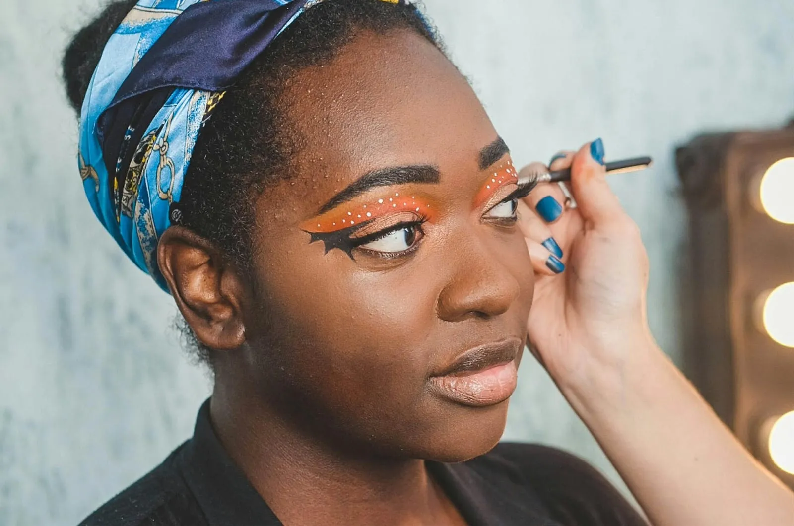 Make-up artist drawing a bat-wing eyeliner flick over orange Halloween eyeshadow dotted with white