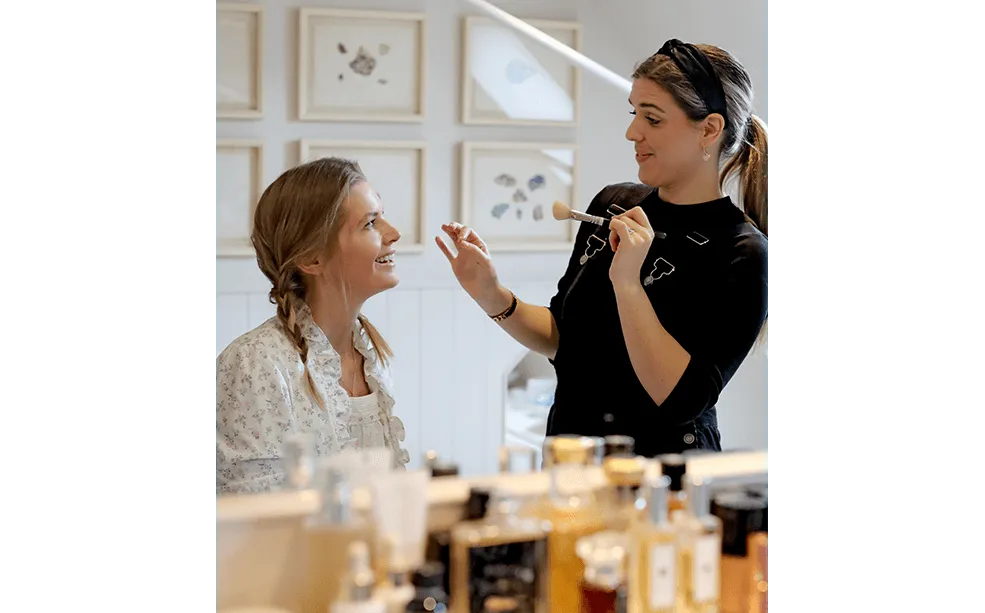Make-up artist holding a brush and chatting to a client at a dressing table lined with perfume bottles