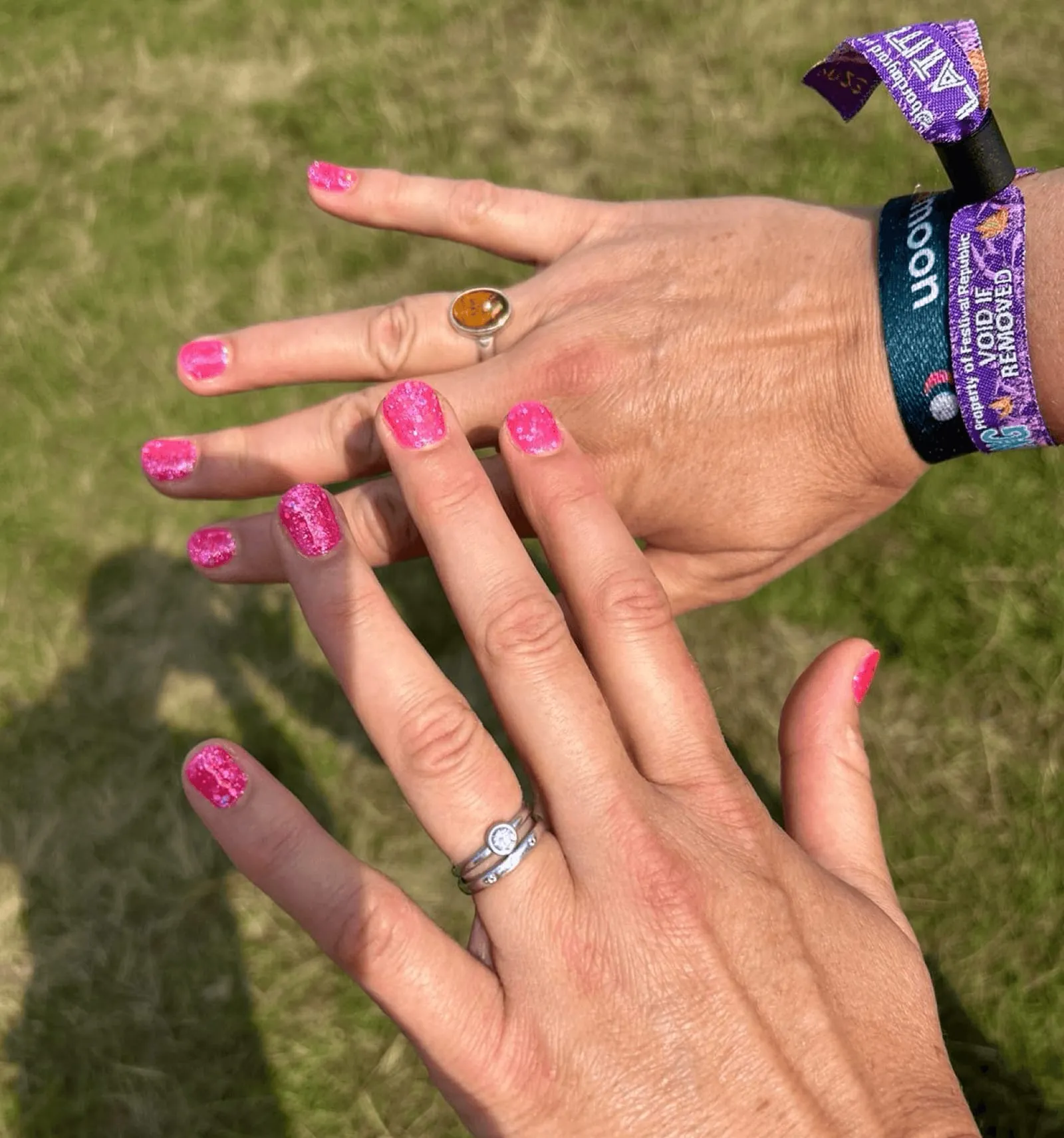 Hands with hot pink glitter gel nails and a festival wristband, held over grass