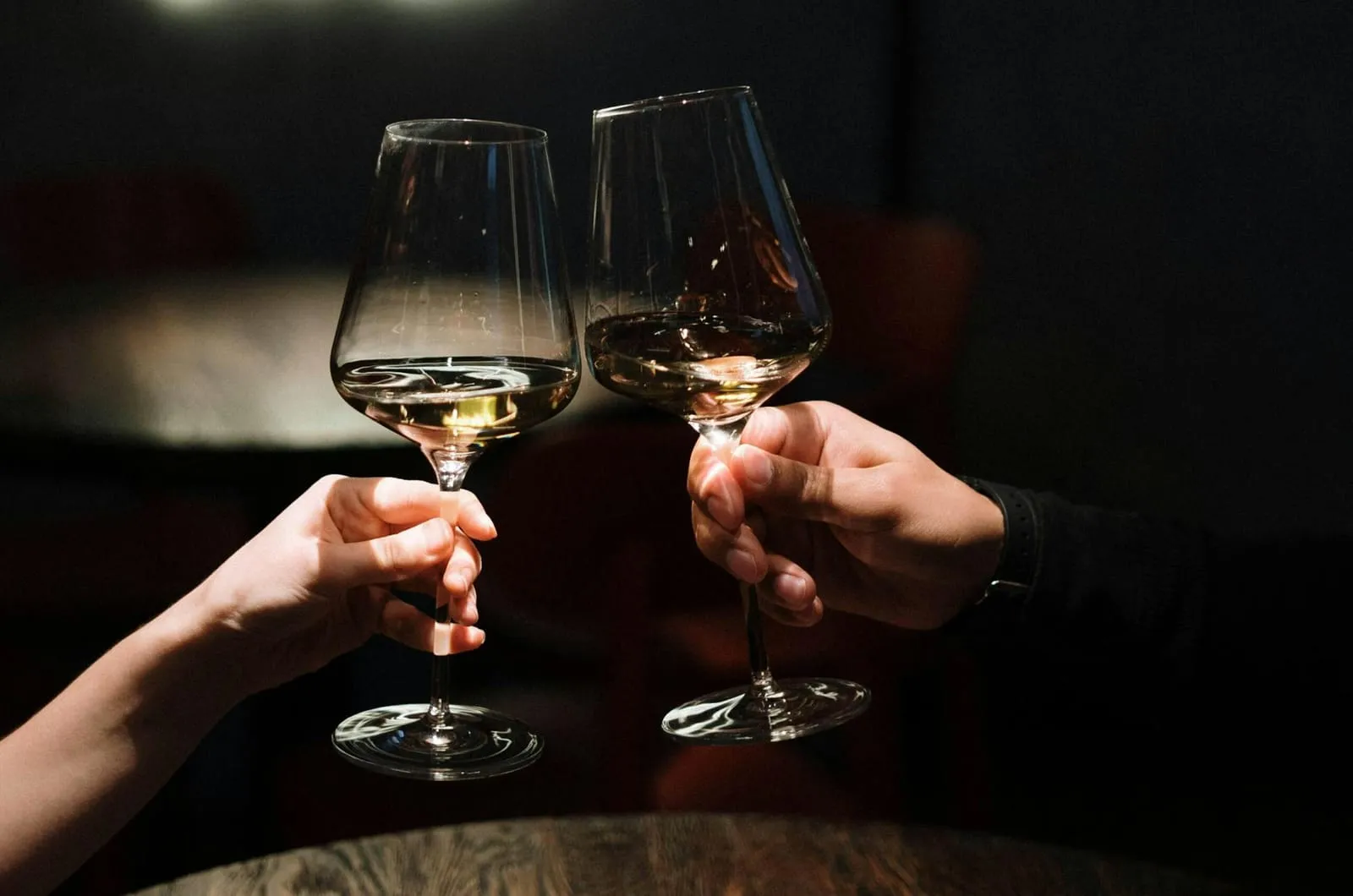 Two people clinking glasses of white wine across a table in a dark, candlelit bar