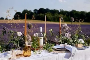 Long outdoor table dressed with tapered candles, foliage and white linen beside a lavender field