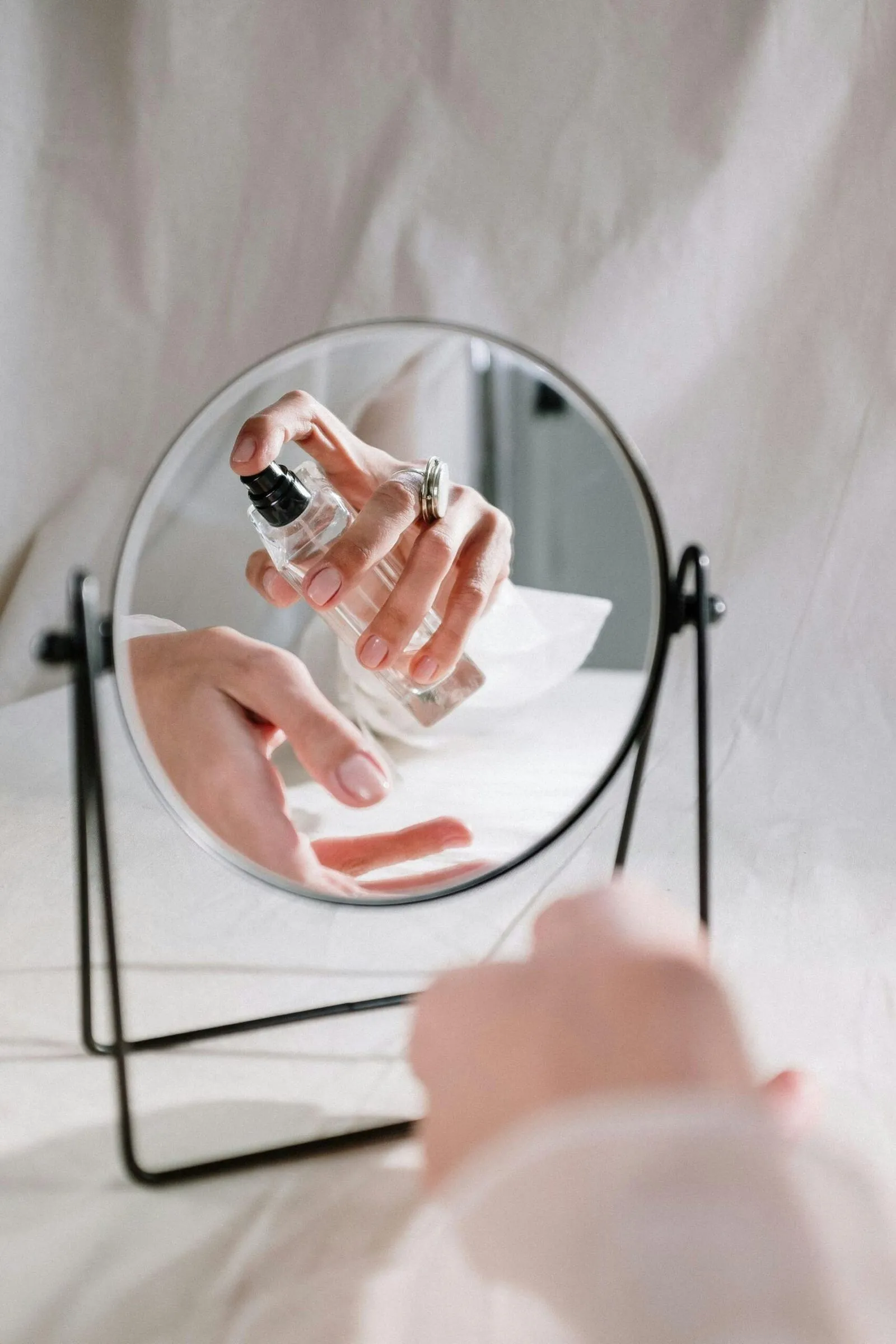 Hand spraying a clear glass perfume bottle, reflected in a round vanity mirror