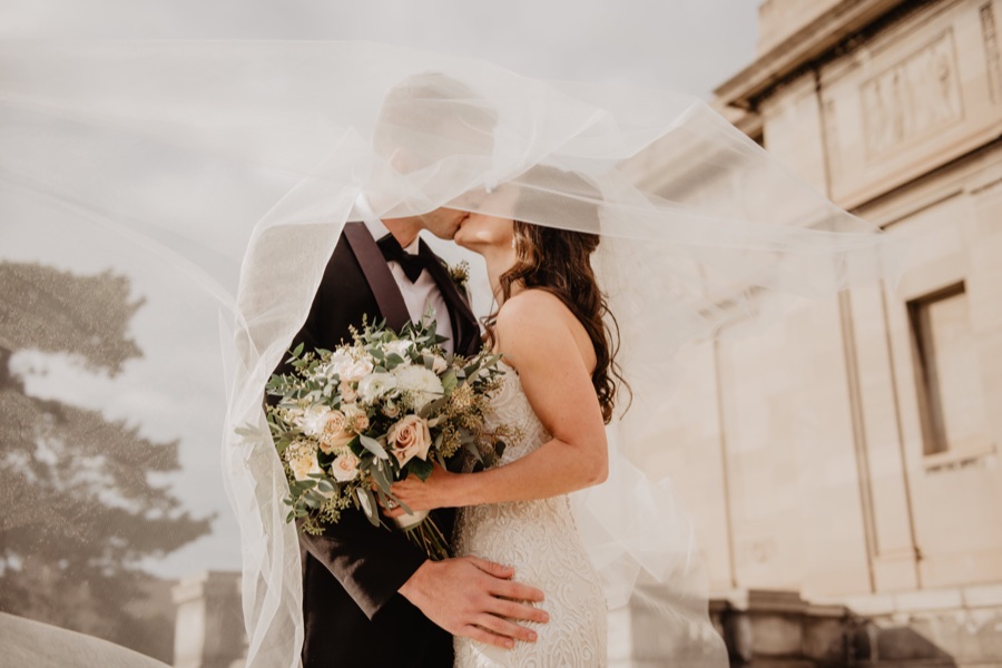 Bride and groom kissing beneath a veil caught in the wind, the bride wearing soft waves and natural bridal make-up